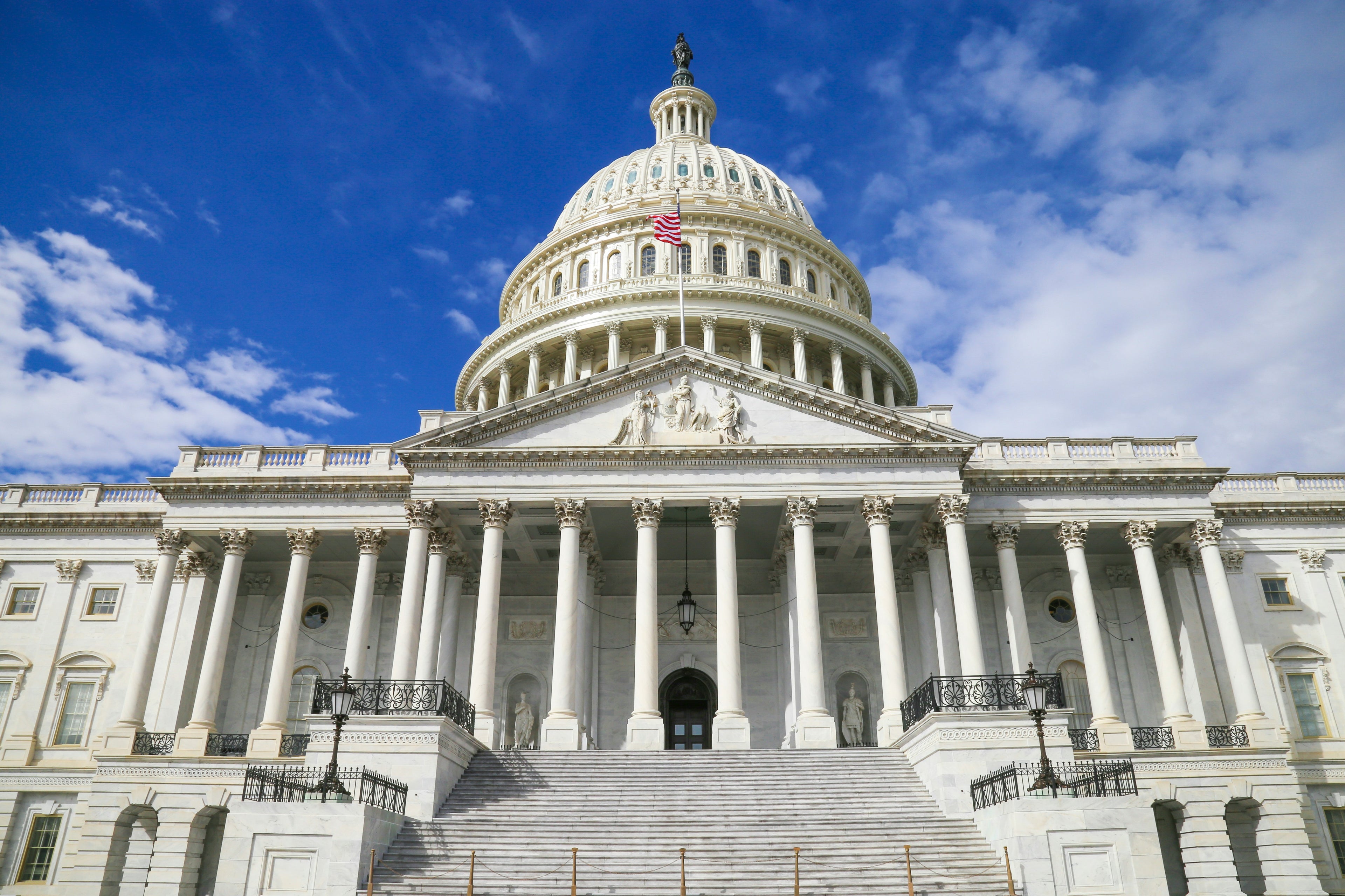 The United States Capitol building with its white neoclassical dome and columns, photographed against a bright blue sky with scattered white clouds.