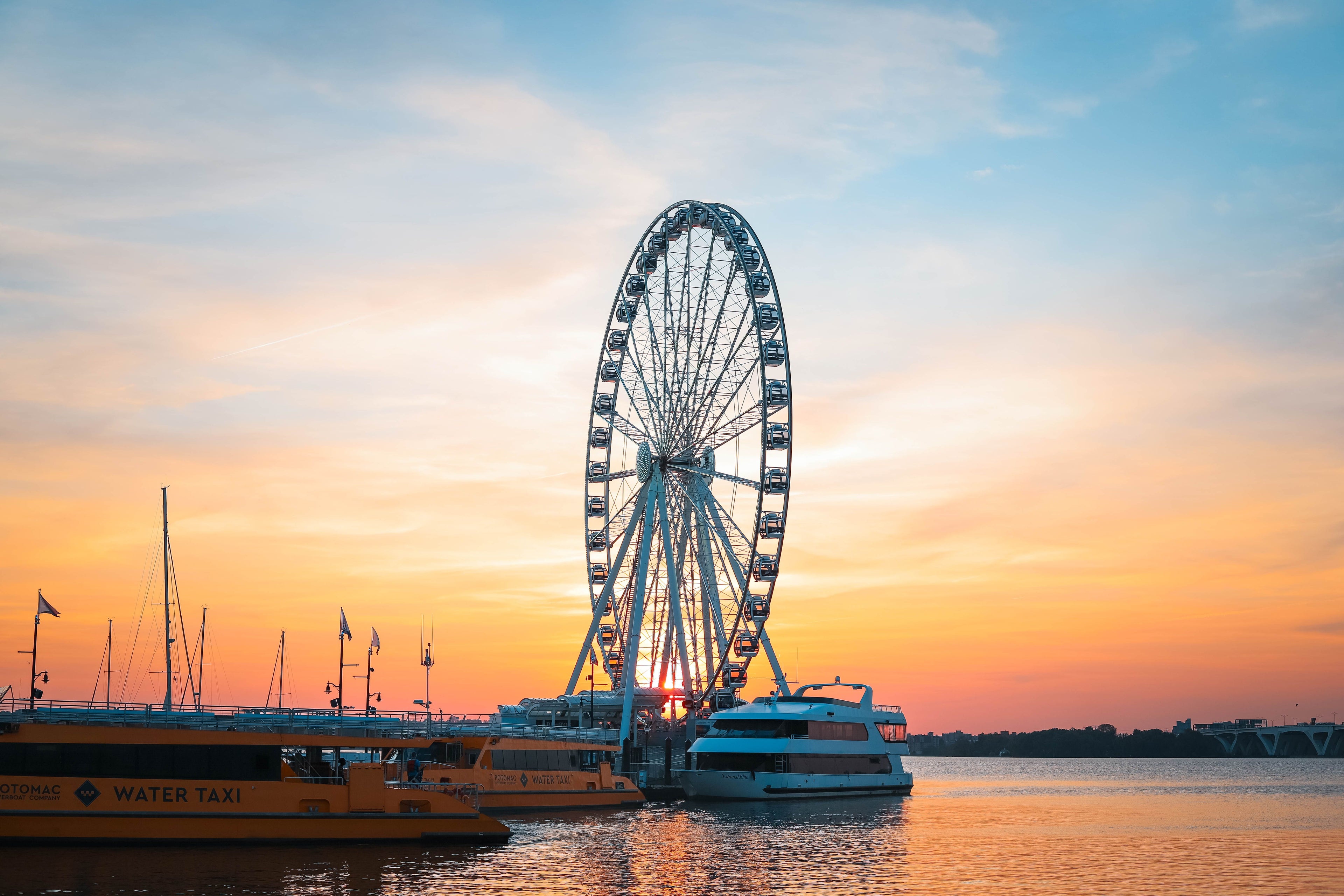 A scenic sunset photograph of a large Ferris wheel over calm water, with boats docked at the pier and a warm orange and blue sky filling the background.