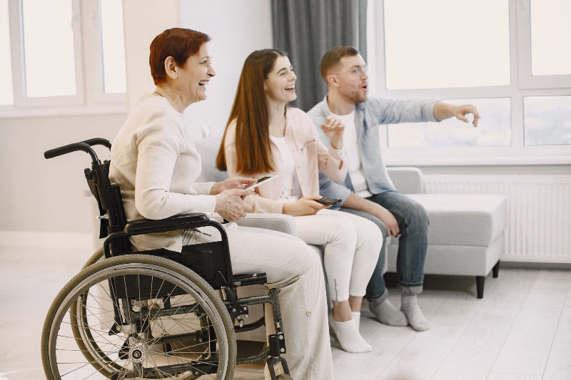 Three people sitting together in a bright living room, with an older woman in a wheelchair rental on the left, a young woman in the center, and a young man on the right pointing toward something outside the window.